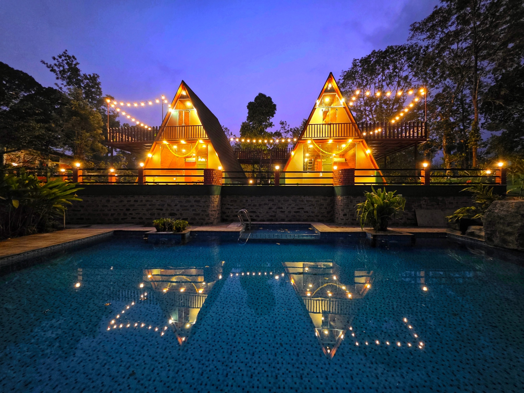 Twin A-frame cottages lit up at dusk beside the swimming pool