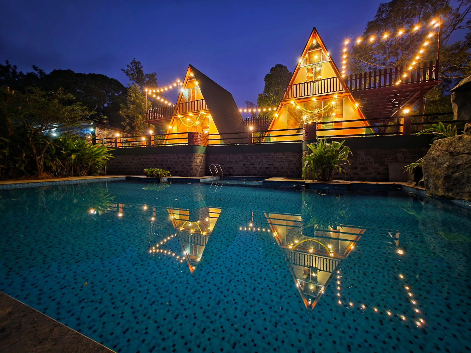 Twin A-Frame cottages lit with fairy lights reflected in the poolside water at dusk